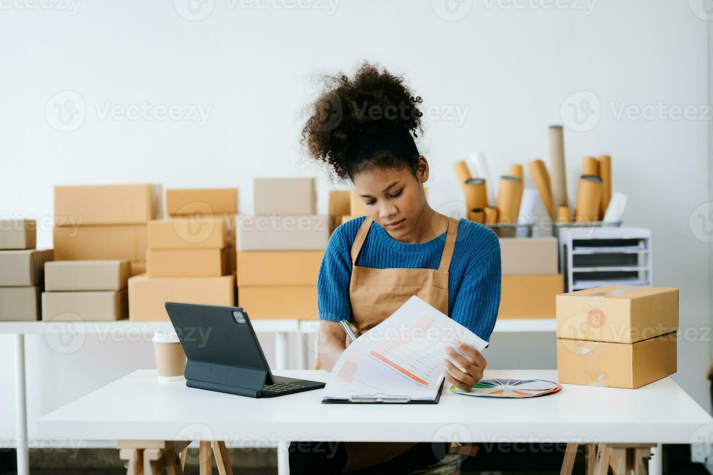 Startup small business SME, Entrepreneur owner African woman using smartphone or tablet taking receive and checking online purchase shopping order to preparing pack product box. in office photo