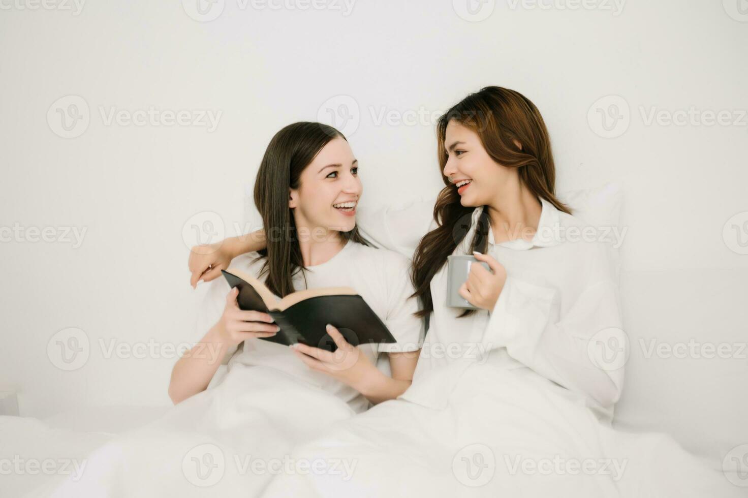 Attractive lesbian couple use mobile phone, tablet watch movie on bed. Beautiful sibling in pajamas lying down with blanket in bedroom together,  in morning photo