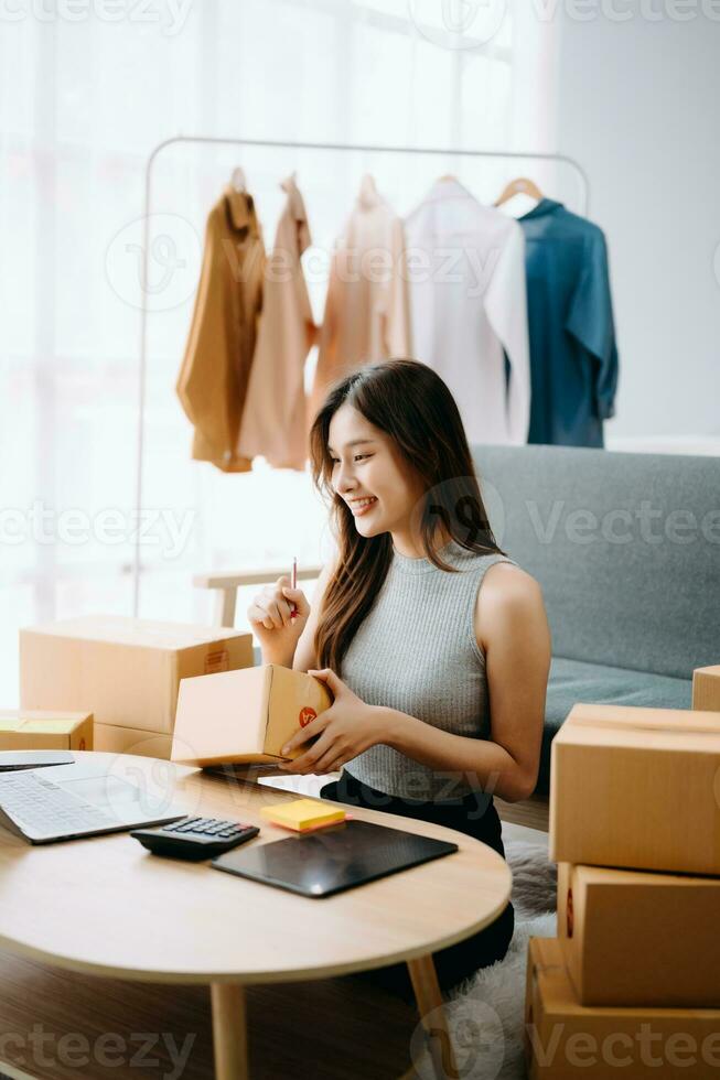 Startup small business SME, Entrepreneur owner woman using smartphone or tablet taking receive and checking online purchase shopping order to preparing pack product box. photo