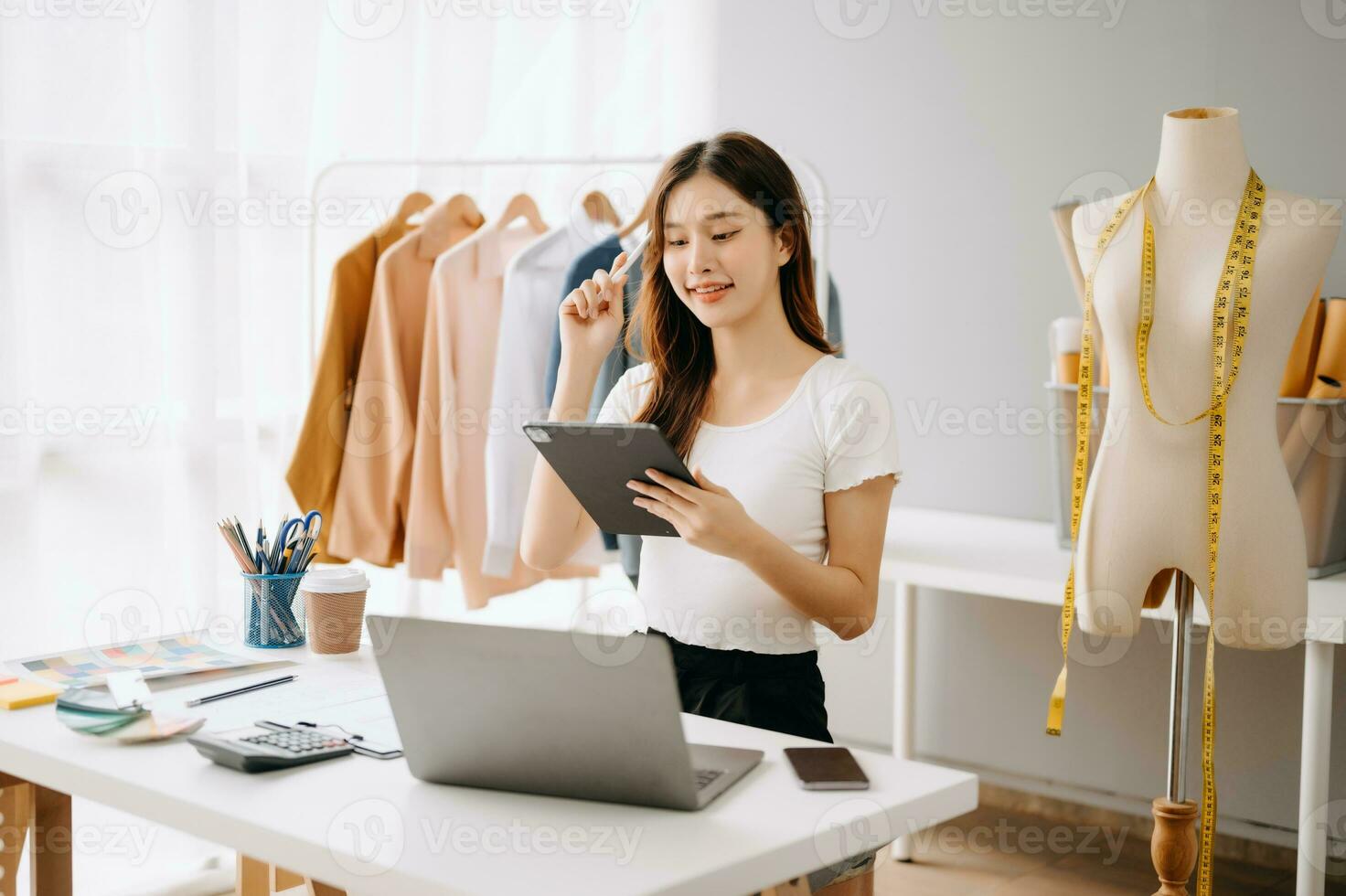 asiático joven mujer en escritorio en oficina de Moda diseñador y sostiene tableta, ordenador portátil y teléfono inteligente en blanco mesa foto