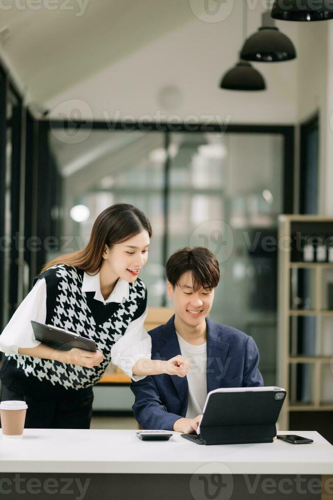 Asian business colleague business presents and explains and using laptop and tablet. Teamwork, financial marketing team, while sitting in modern office room. photo