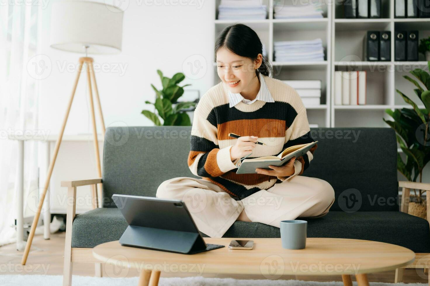 Attractive happy young Asian student studying at the college library, sitting at sofa, using a laptop computer, tablet and headphones having a video chat. photo