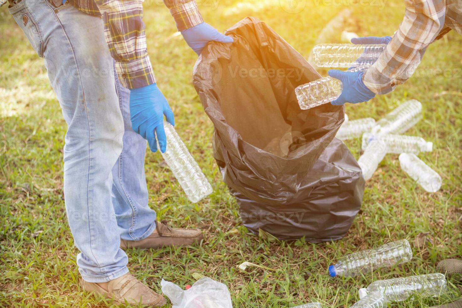Two man employees use black garbage bags to collect plastic bottles and recyclable waste from the lawn and sidewalks for recycling. Concept of sorting plastic waste for recycling photo