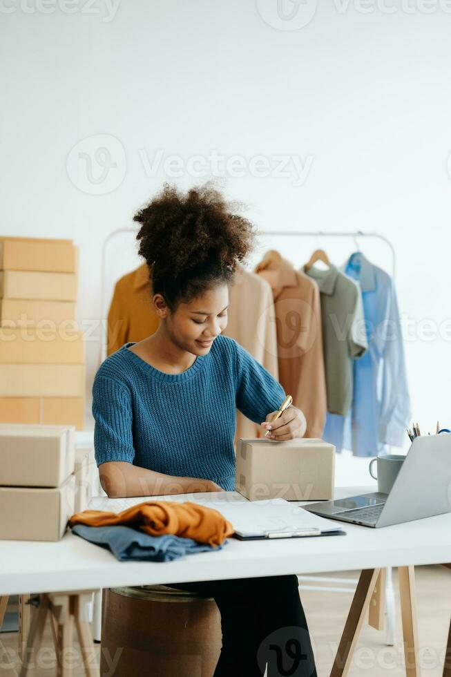 Young African woman running online store Startup small business SME, using smartphone or tablet taking receive and checking online purchase shopping order in office photo