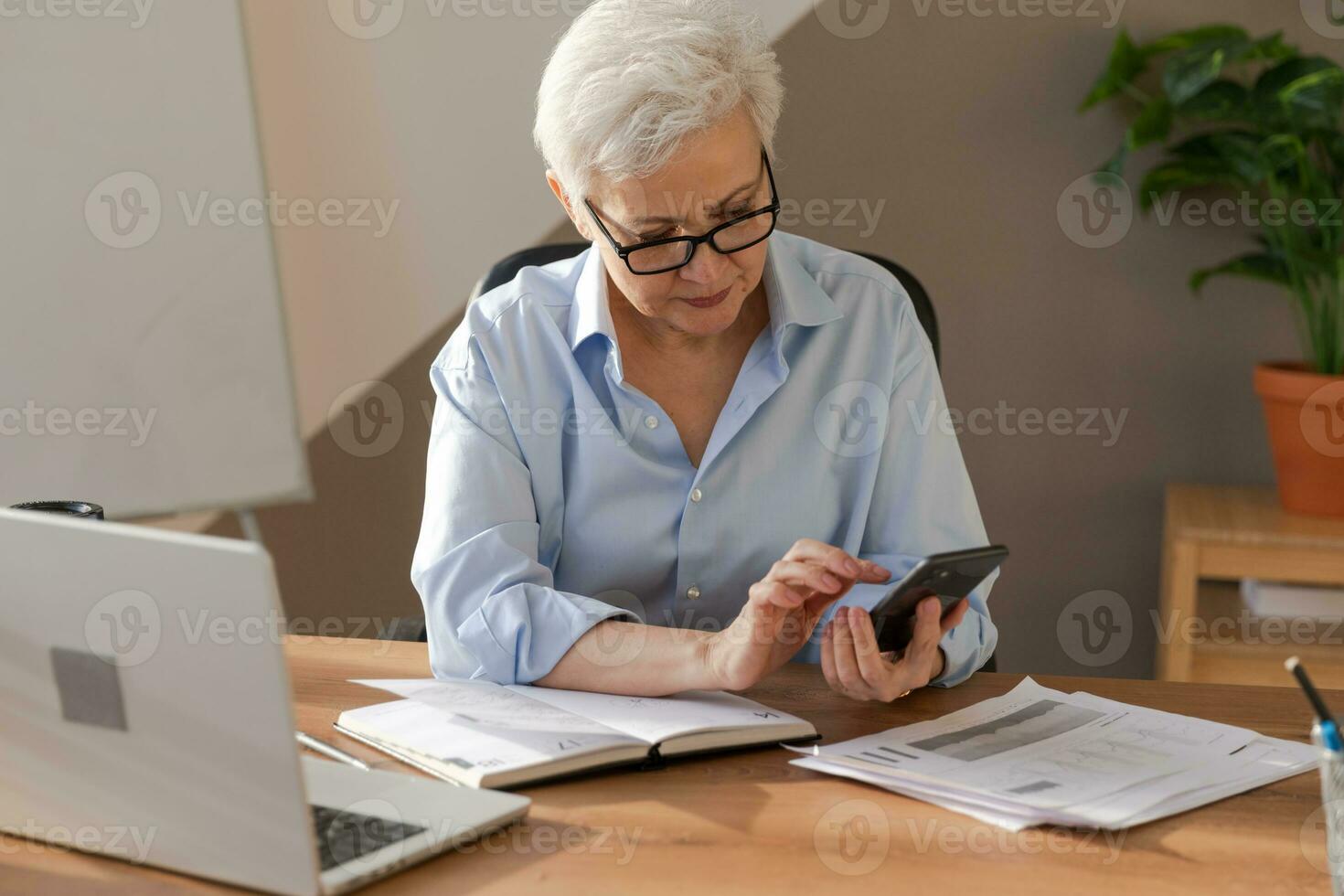 Confident stylish european middle aged senior woman using smartphone at workplace. Stylish older mature 60s gray haired lady businesswoman with cell phone in office. Boss leader using internet apps. photo