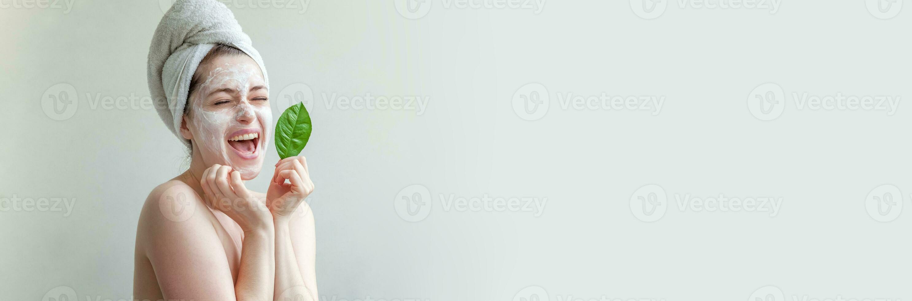 Beauty portrait of woman in towel on head with white nourishing mask or creme on face and green leaf in hand, white background isolated. Skincare cleansing eco organic cosmetic spa concept. photo