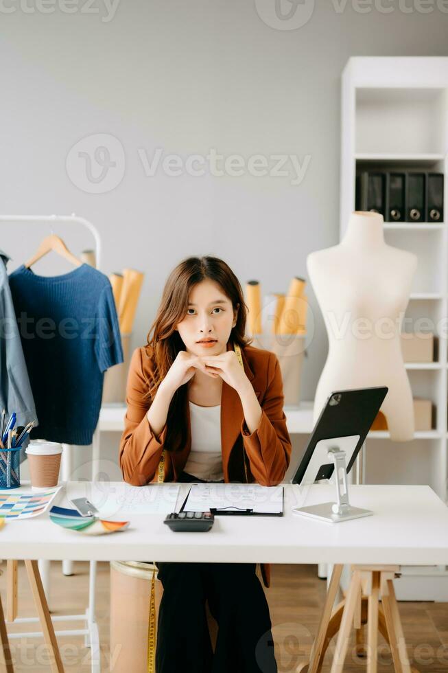 Asian tailor woman working on clothes in tailoring atelier.  beautiful young female fashion designer smile and after success in studio photo