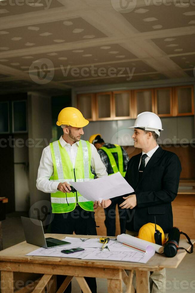 Contractor and architect looking at plan, tablet, laptop and talking about working on big project. Building in construction interior. photo