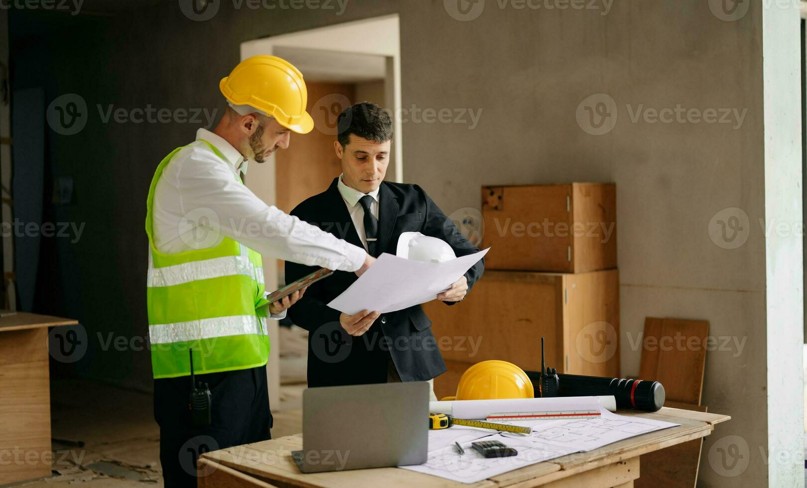 Contractor and architect looking at plan, tablet, laptop and talking about working on big project. Building in construction interior. photo