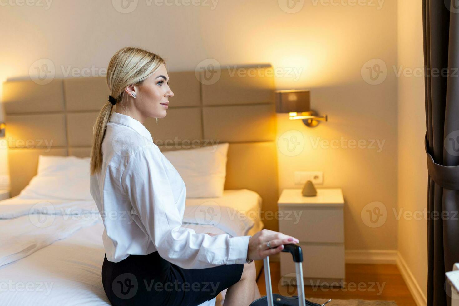 Young businesswoman sitting on bed holding suitcase in hotel room. Business woman in suit with luggage in hotel room. Business trip photo