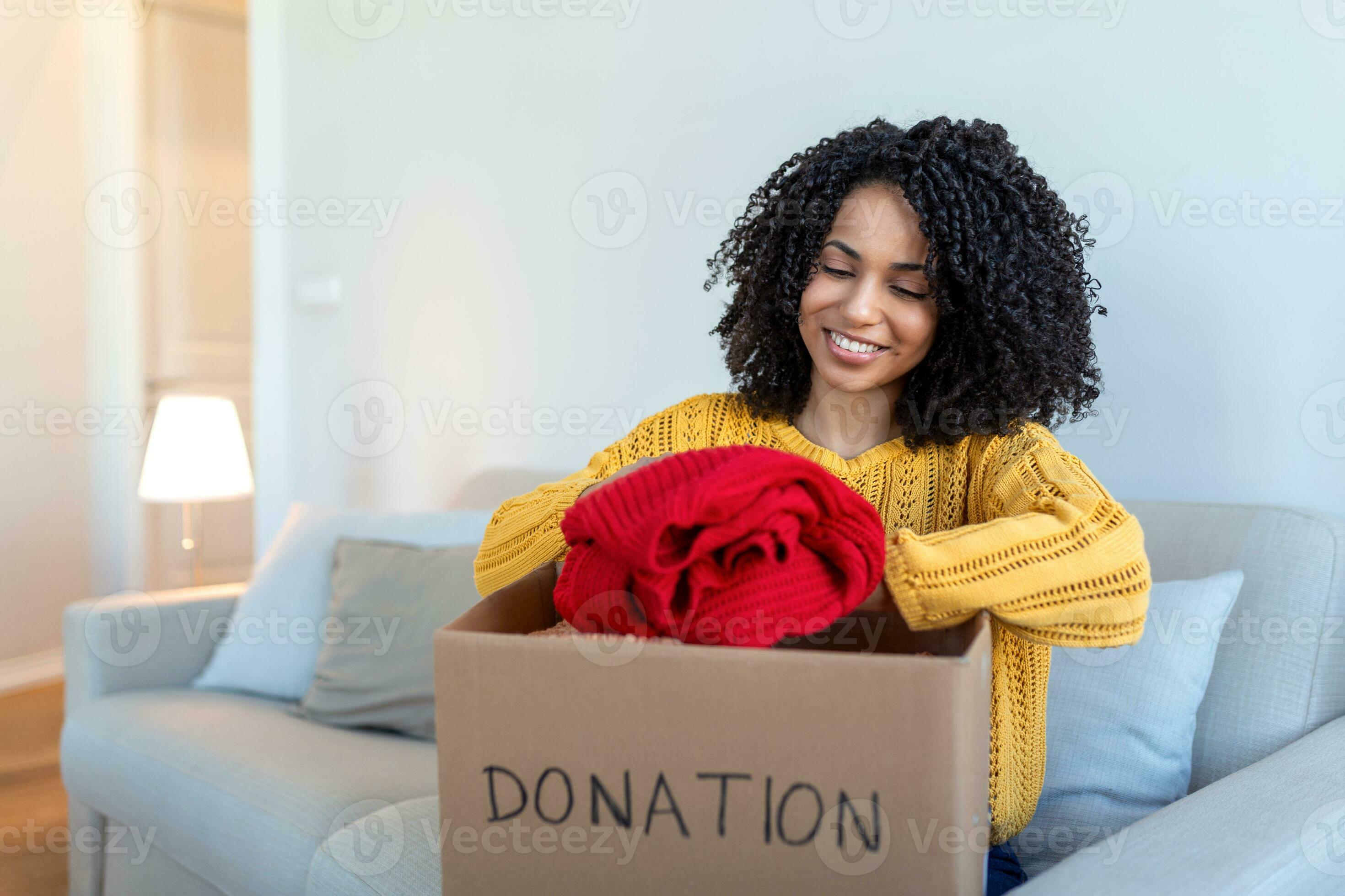 Happy African young woman sit on couch stuck clothes in donation box at