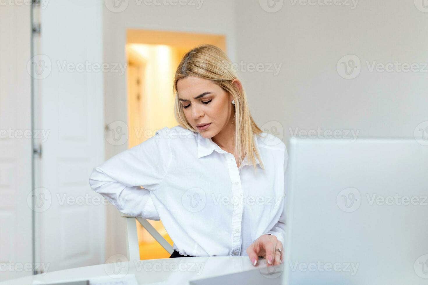 Portrait of young stressed woman sitting at home office desk in front of laptop, touching aching back with pained expression, suffering from backache after working on pc photo