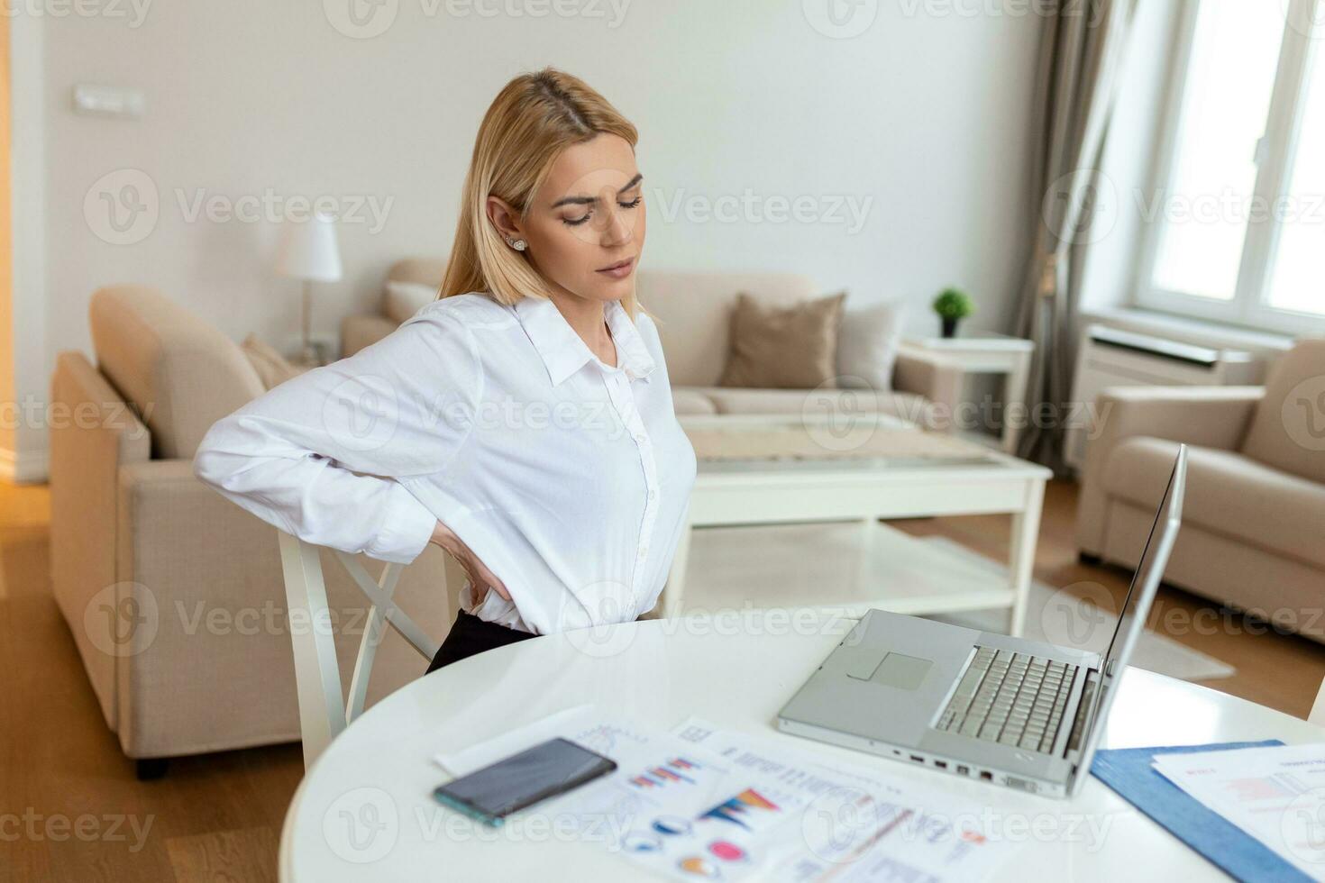 Portrait of young stressed woman sitting at home office desk in front of laptop, touching aching back with pained expression, suffering from backache after working on pc photo