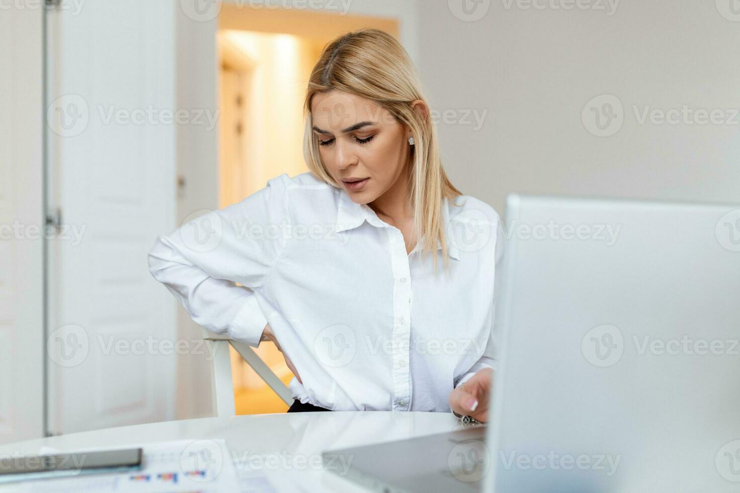 Portrait of young stressed woman sitting at home office desk in front of laptop, touching aching back with pained expression, suffering from backache after working on pc photo