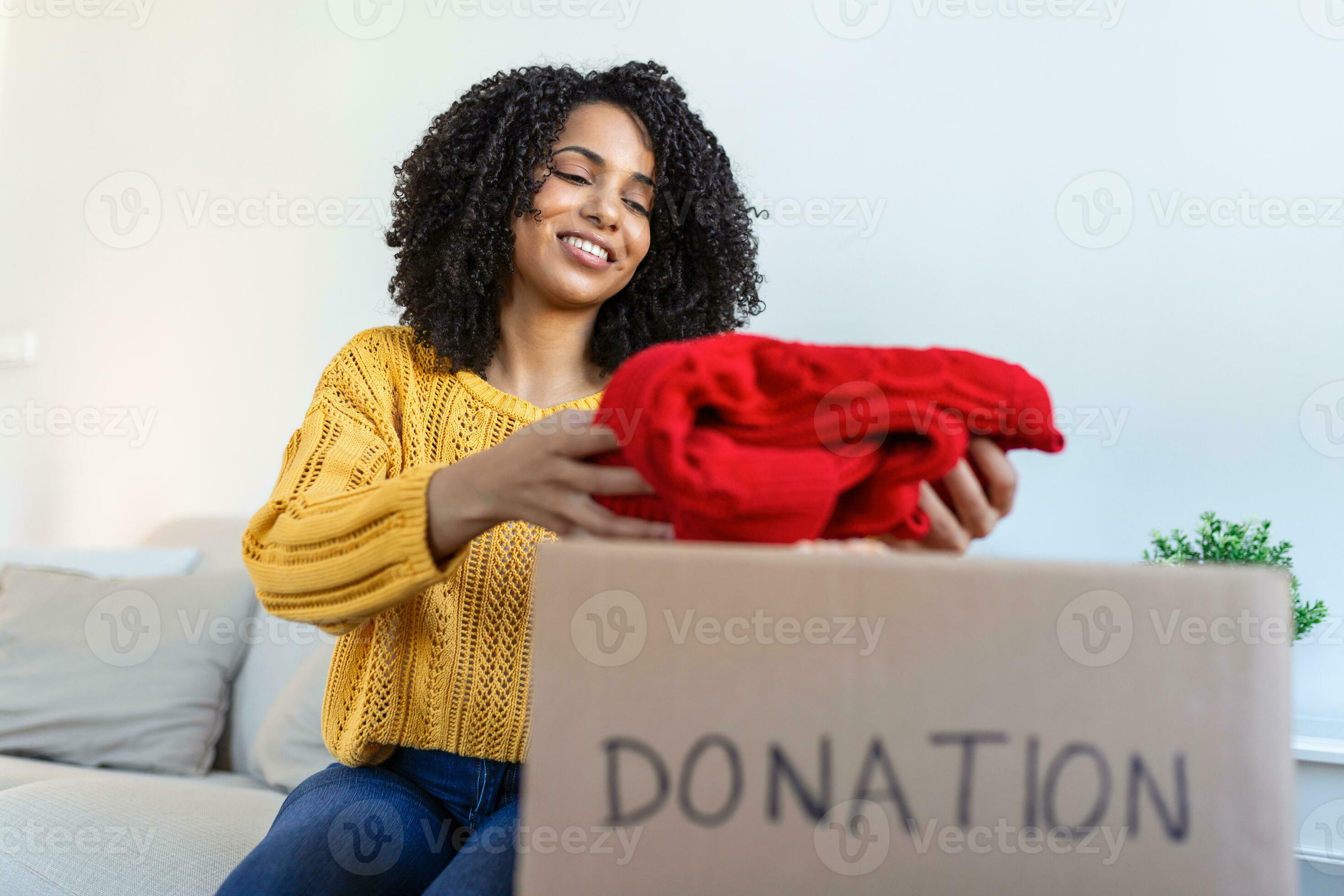 Happy African young woman sit on couch stuck clothes in donation box at