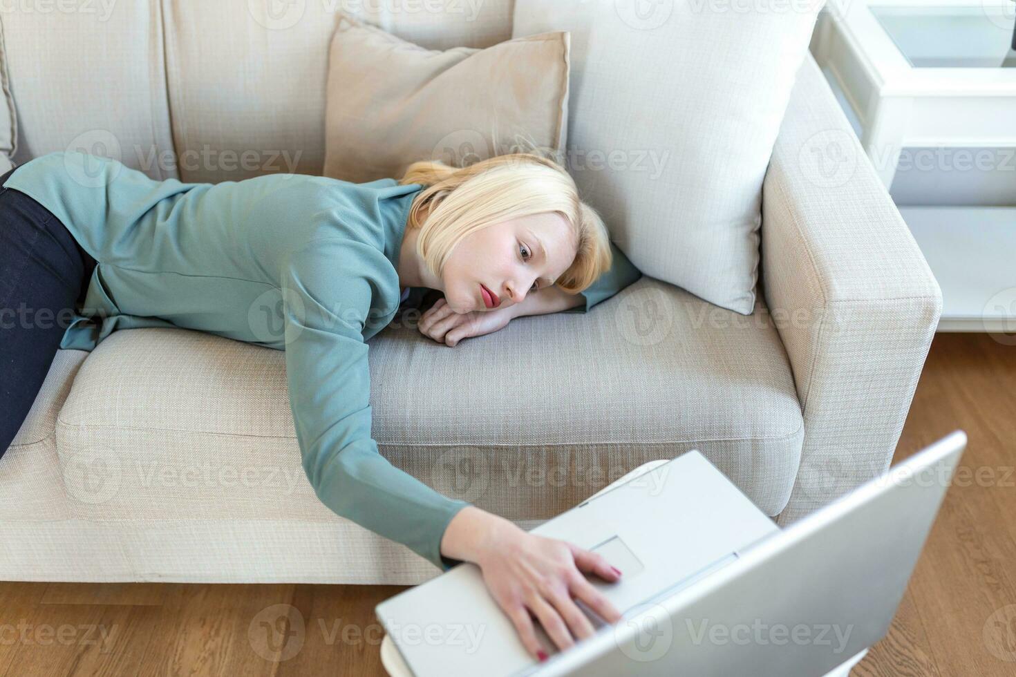 Exhausted young woman lying on sofa, using laptop, too tired or bored of online work at home, free space. Workaholism, chronic fatigue, overworking on remote job concept photo
