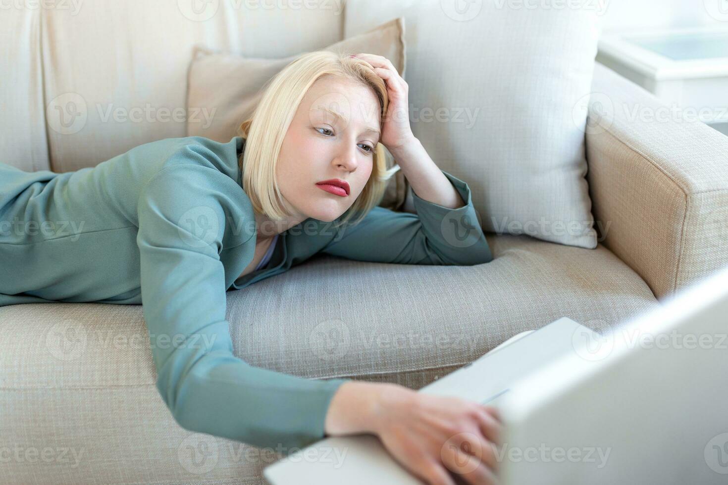 Exhausted young woman lying on sofa, using laptop, too tired or bored of online work at home, free space. Workaholism, chronic fatigue, overworking on remote job concept photo