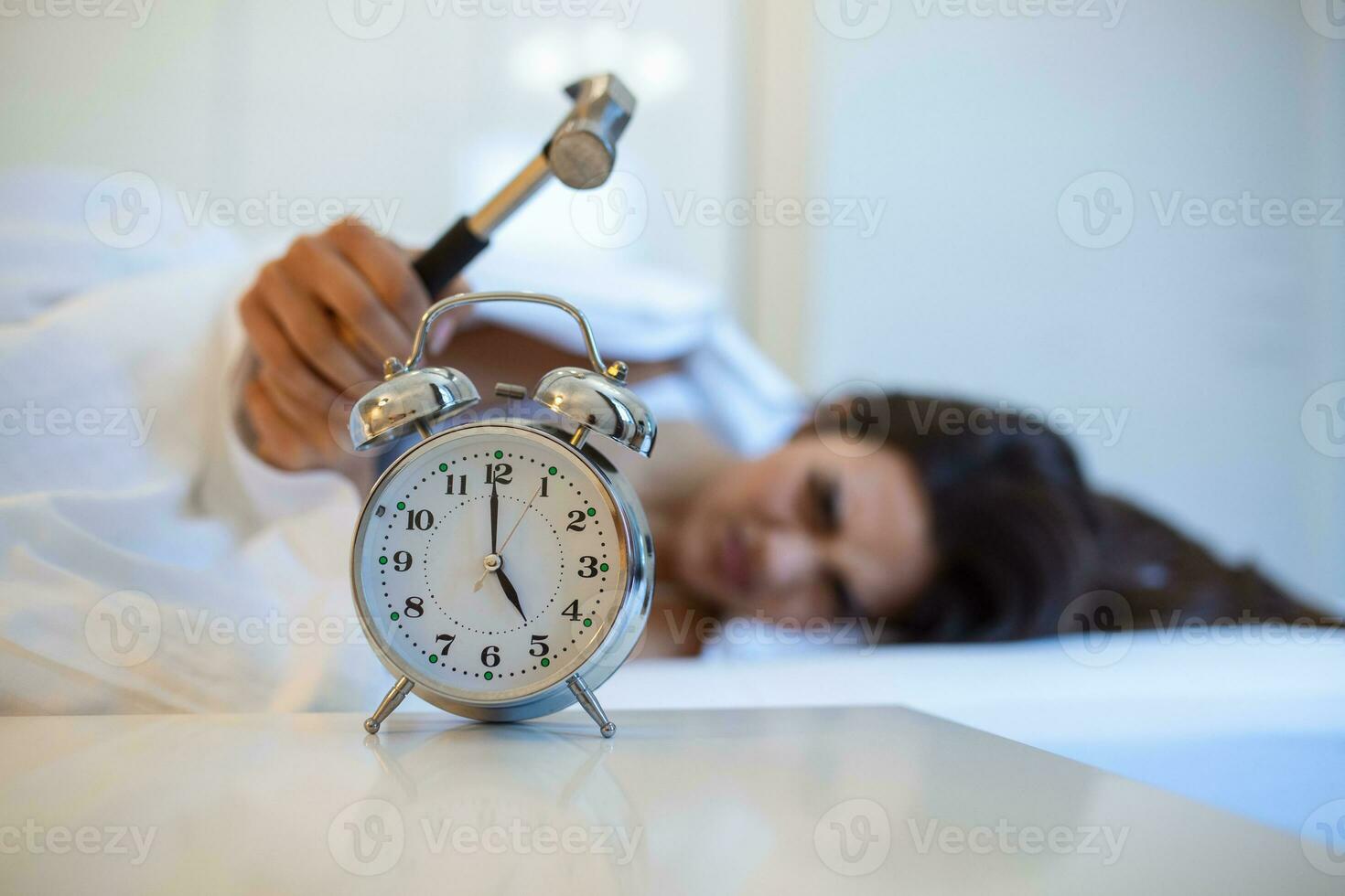 Young woman tries to break the alarm clock with hammer, Destroy the Clock. Girl lying in bed turning off an alarm clock with hammer in the morning at 5am. photo