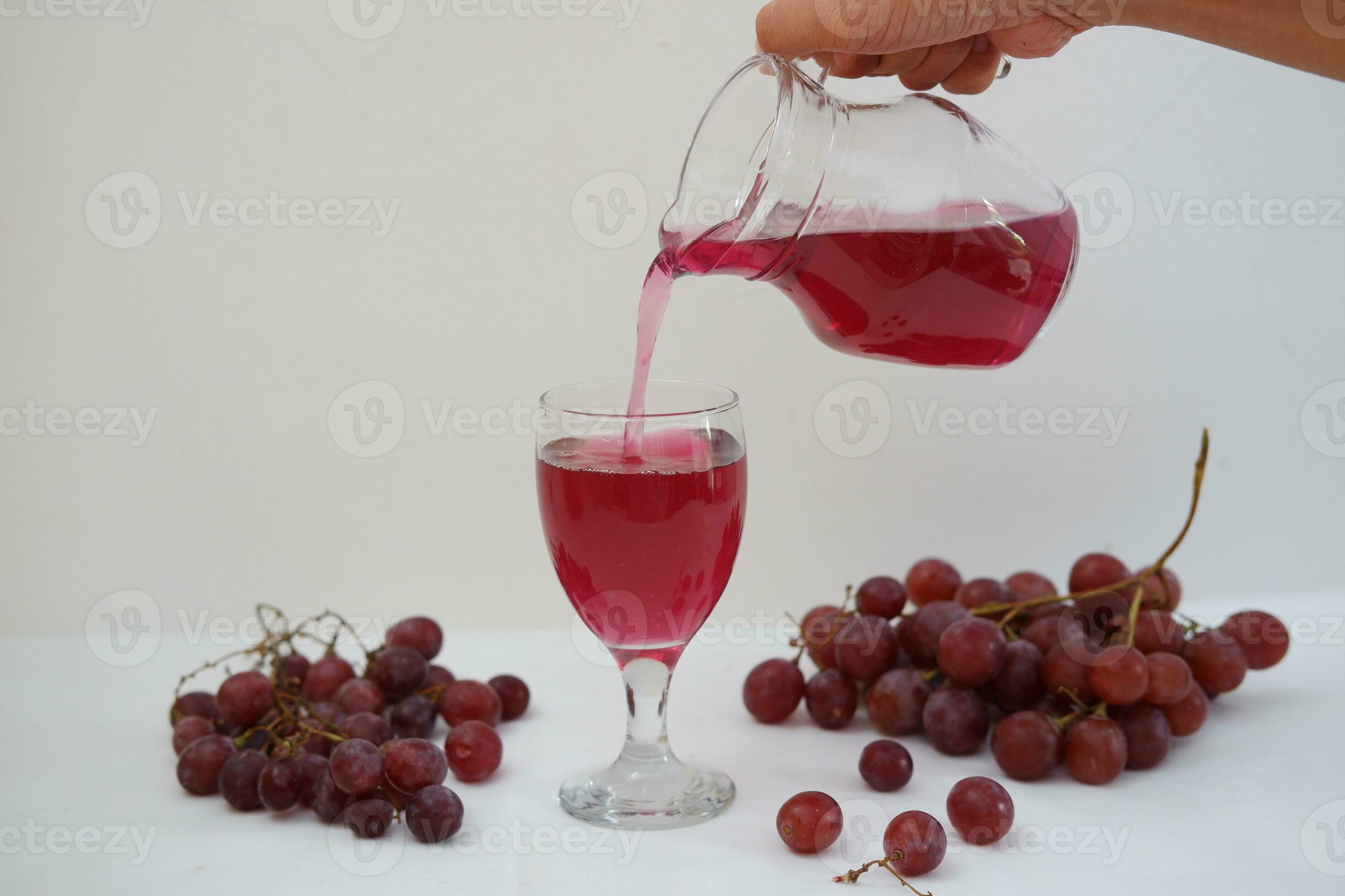 Red grape juice pouring into a glass with fresh grapes on white