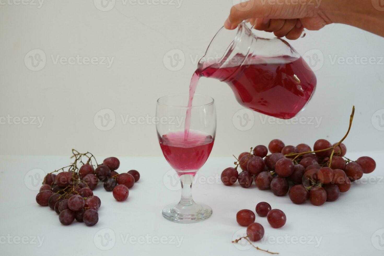 Red grape juice pouring into a glass with fresh grapes on white