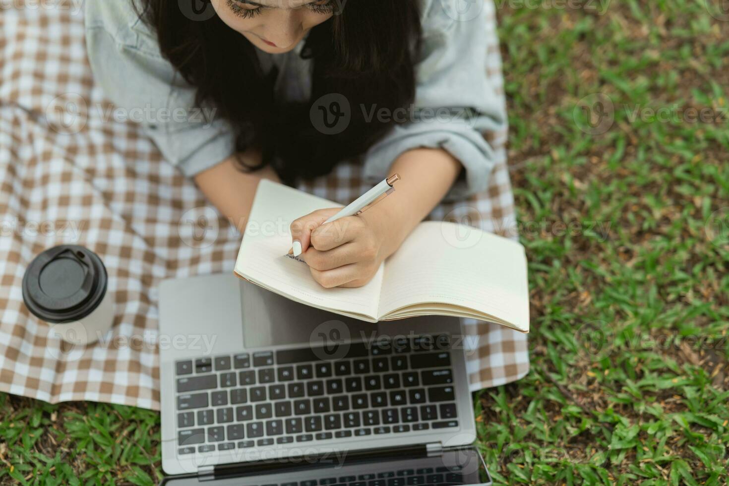 Top view. Asian woman lay on picnic blanket and lawn at park garden working on laptop and writing on notebook. Asian female using laptop while sitting under a tree at park. Work from anywhere concept. photo