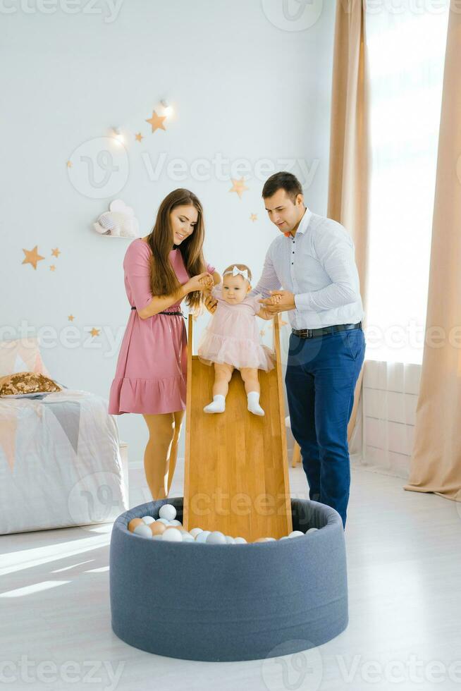 joven hermosa caucásico familia jugando con su un año de edad hija en el para niños habitación. el niña diapositivas abajo el diapositiva dentro el piscina con pelotas foto