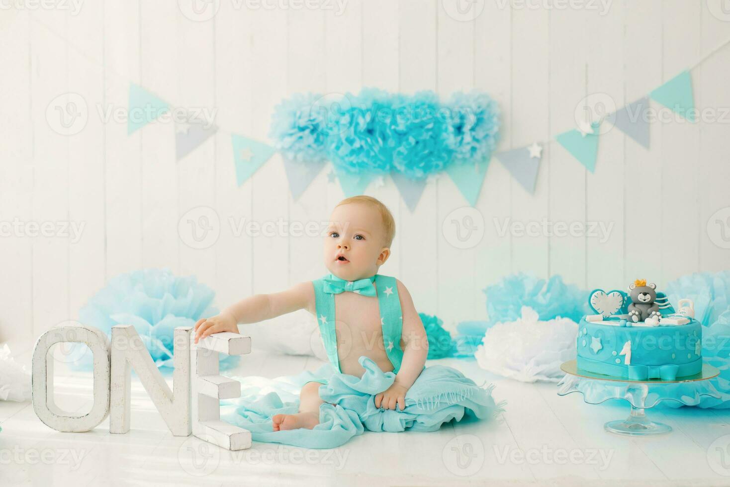 A one-year-old boy in blue shorts and a bow tie celebrates his birthday next to the letters ONE on a stand and a decoration for his party photo