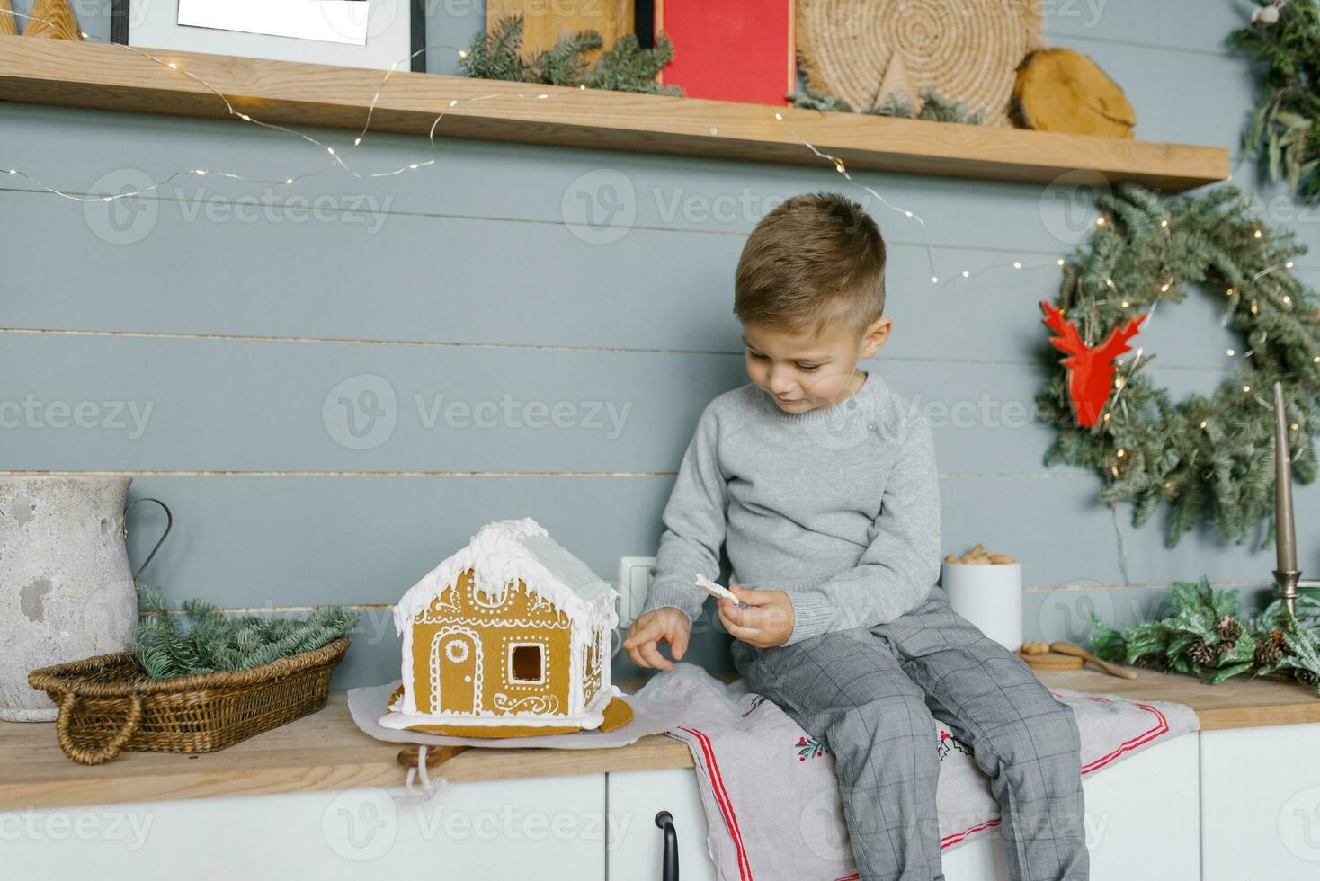 A beautiful baby boy is sitting on the kitchen table and decorating a gingerbread house for Christmas photo