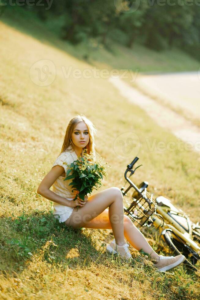 portrait of a beautiful girl in a field, sitting on the grass, next to a bicycle, with a bouquet of verbena flowers in the setting sun in summer photo