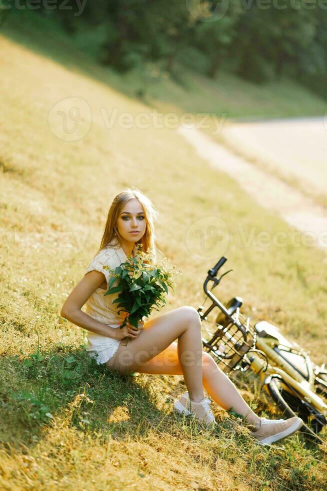 retrato de un hermosa niña en un campo, sentado en el césped, siguiente a un bicicleta, con un ramo de flores de verbena flores en el ajuste Dom en verano foto