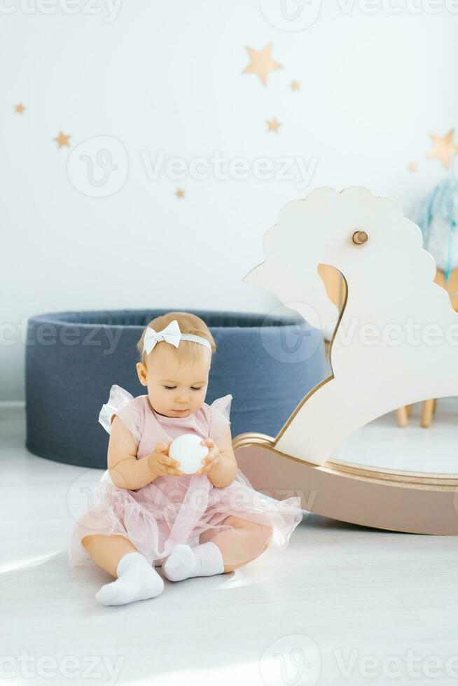 A cute blue-eyed one-year-old baby in a pink dress holds a small white ball in her hands and sits on the floor in the children's room photo