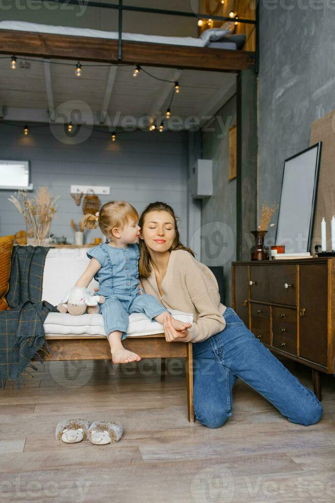 dos años de edad hija Besos su madre en el mejilla, sentado en el sofá foto