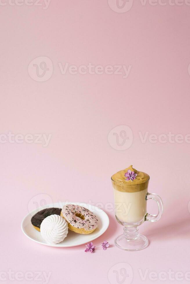 Beautiful dalgon coffee drink. A Cup of coffee with foam and lilac flower, a plate of sweets, marshmallows and donuts on a pink background. photo