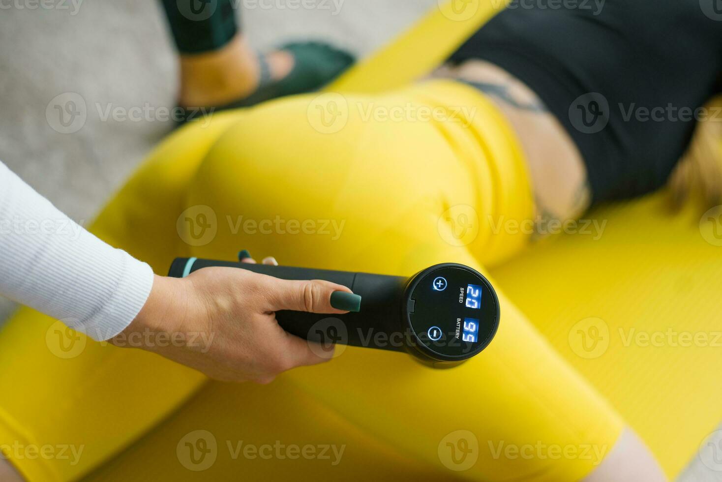 Female massage therapist does massage exercises on the hips with the help of a shock vibrator massager to a female client at home. Shock therapy for a restorative massage of the athletic body photo