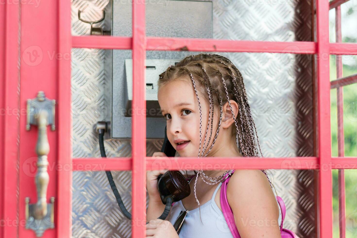 un sonriente niña con trenzas afro hace un teléfono llamada en un antiguo Inglés teléfono puesto. generacional contraste foto