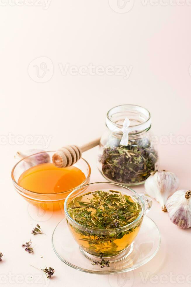 Hot tea with oregano in a cup, honey in a bowl, heads of garlic and dry herb in a jar. Herbal medicine and alternative therapy. Vertical view photo