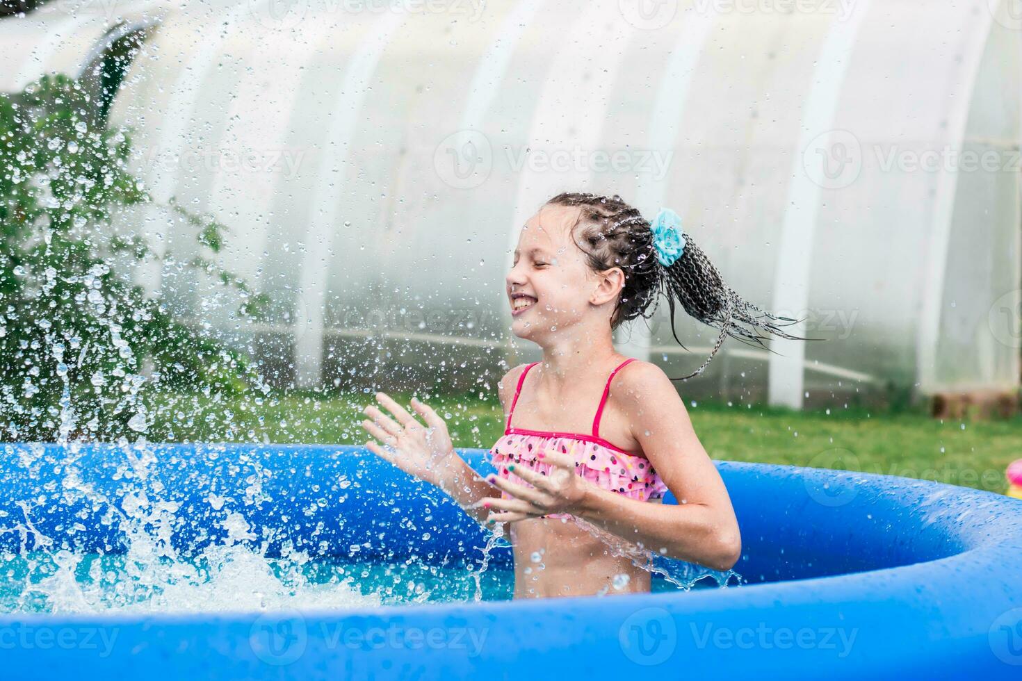 alegre pequeño niña con trenzas afro salpicaduras agua en un inflable piscina en un verano día en el patio interior foto