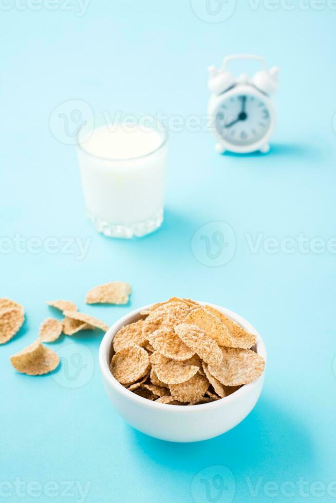 A bowl of cereal, a glass of milk and an alarm clock on a blue background. Scheduled breakfast. Vertical view photo