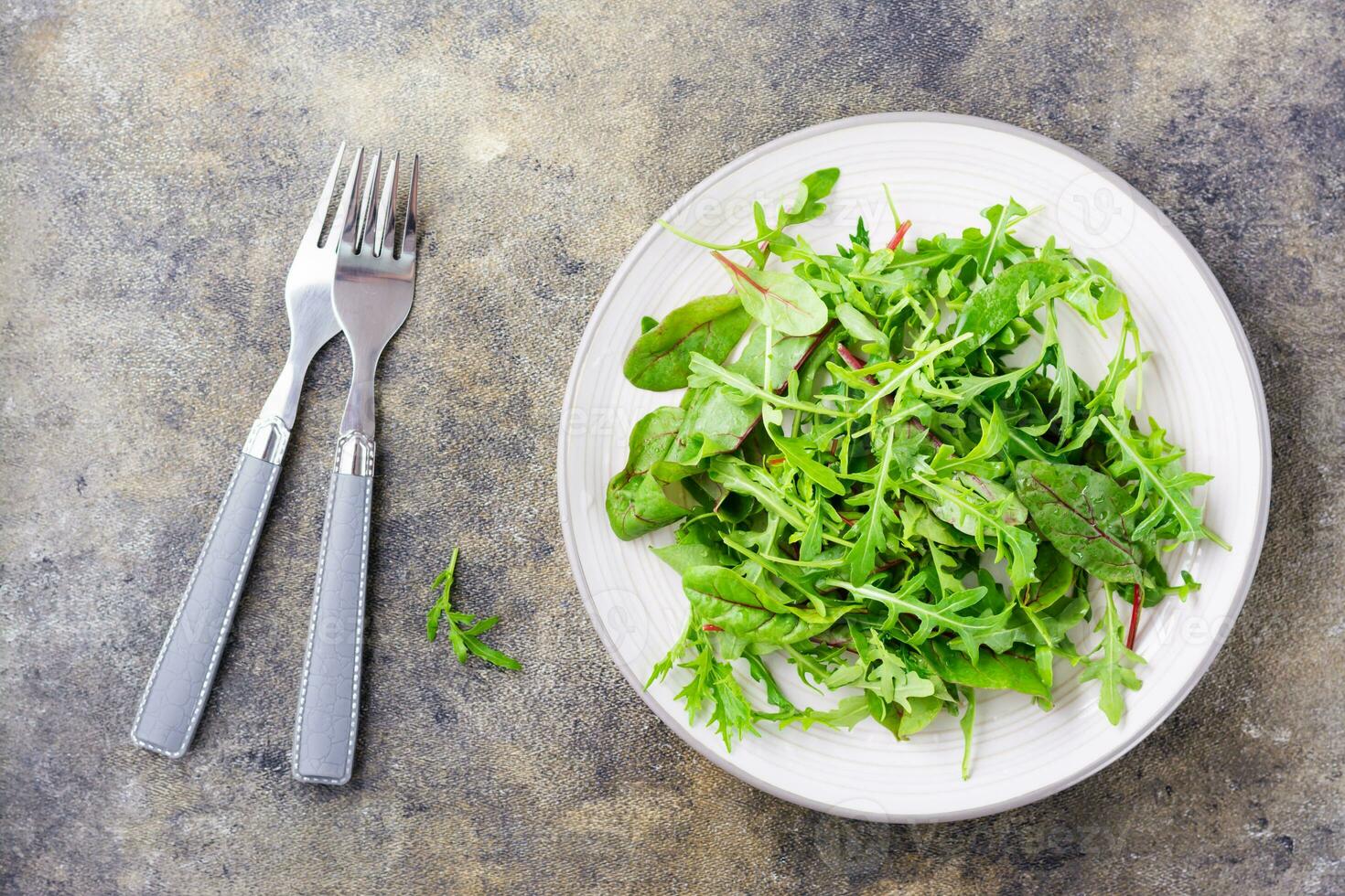A mixture of fresh arugula, chard and mizun leaves on a plate and forks on the table. Vegetarianism, healthy eating. Top view photo