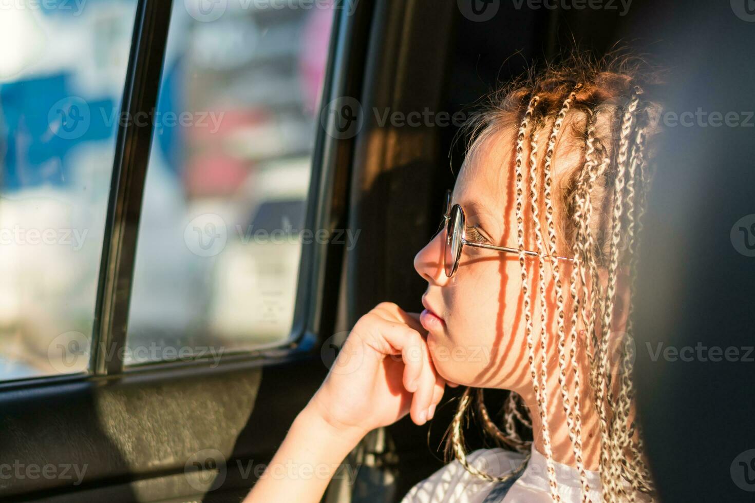 A pensive teenager girl with afro-braids and sunglasses looks out the window while sitting in the car. Road trip photo