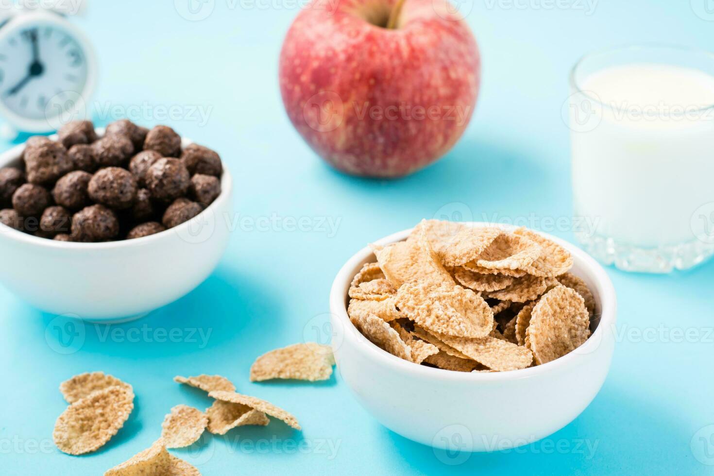 Bowls with cereal and chocolate balls, a glass of milk, an apple and an alarm clock on a blue background. Scheduled breakfast. Close-up photo