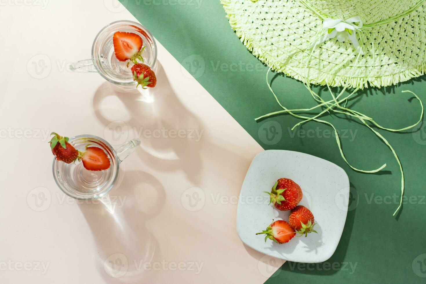 Infused water with strawberries in glasses, berries on a saucer and a straw hat on a green-pink background in harsh light with shadows. Detox drink. Top view photo