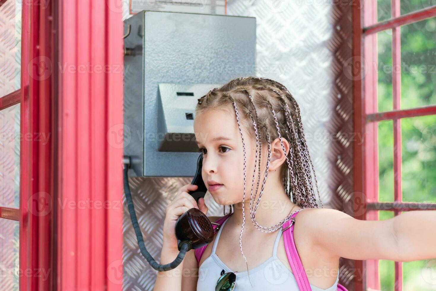 un grave niña con trenzas afro hace un teléfono llamada en un antiguo Inglés teléfono puesto. generacional contraste foto