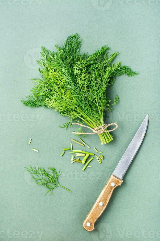 A knife cuts off the stems from a bunch of fresh dill on a green background. Vitamin greens in a healthy diet. Top and vertical view photo