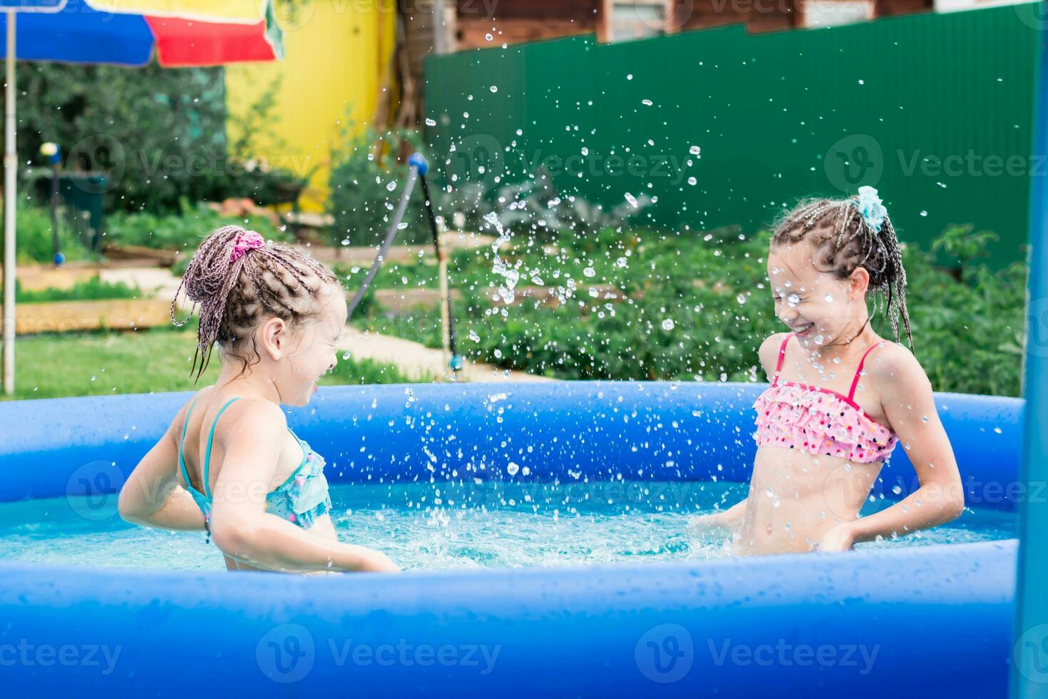 Two girls with afro-braids have fun splashing water in an inflatable pool on a summer day in the backyard photo