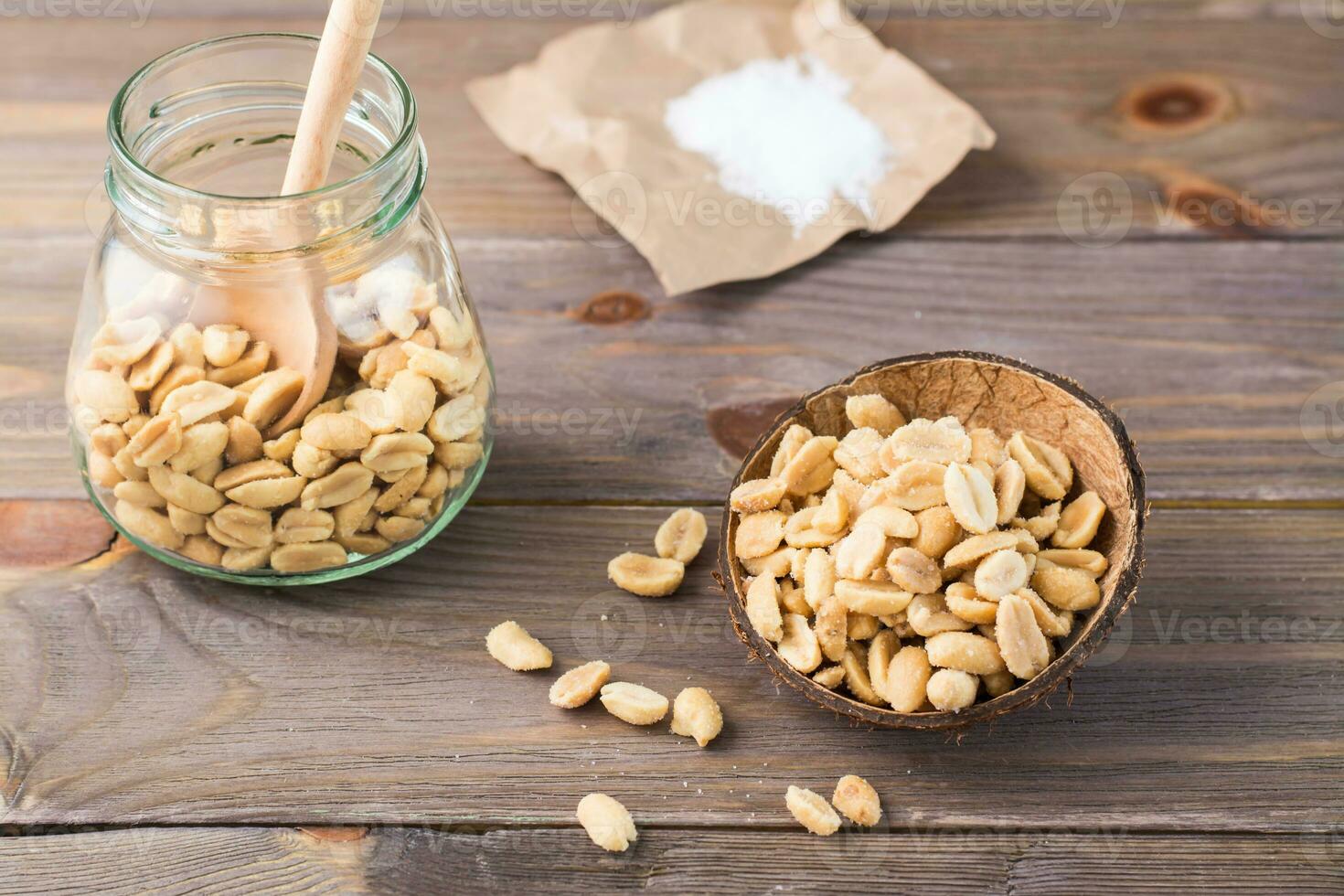 Salted peanuts in a bowl and in a jar with a spoon and salt on paper on a wooden table. Healthy vegetarian food. Rustic style photo