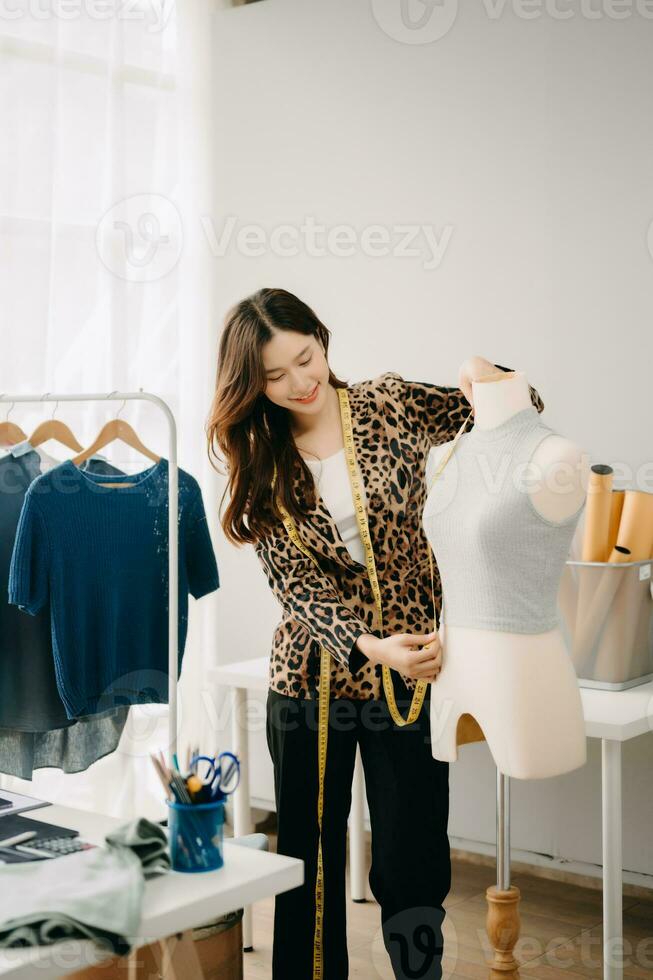 Successful fashion designer. Attractive young asian woman with smile while standing in workshop. Beautiful owner business woman working and holds tablet, laptop and smartphone on desk in studio. photo