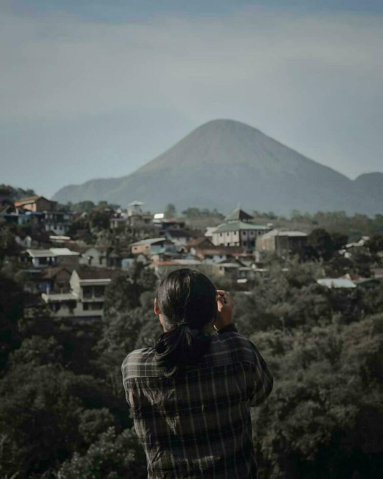 Individual in Rear View, Surrounded by Landscape and Mountains Adult admiring mountain range with buildings, crowd, and blue sky. photo