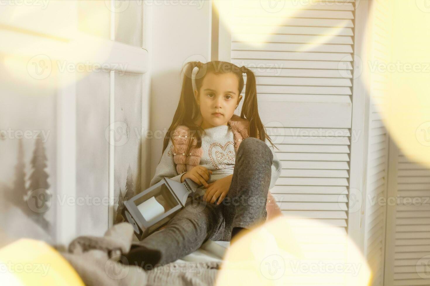 little girl sitting by the window in the children's room during Christmas time, preparing to celebrate Xmas Eve, view through a window from outside into a decorated lights photo