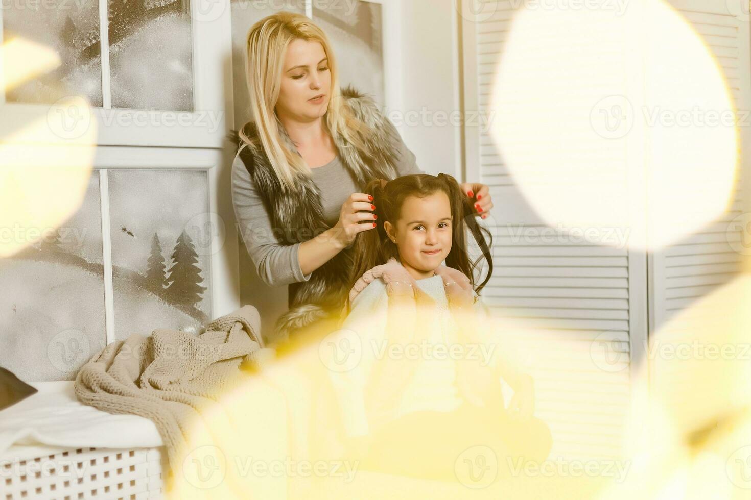 contento amoroso familia. madre es peinada su hija pelo sentado por el ventana en el para niños habitación foto